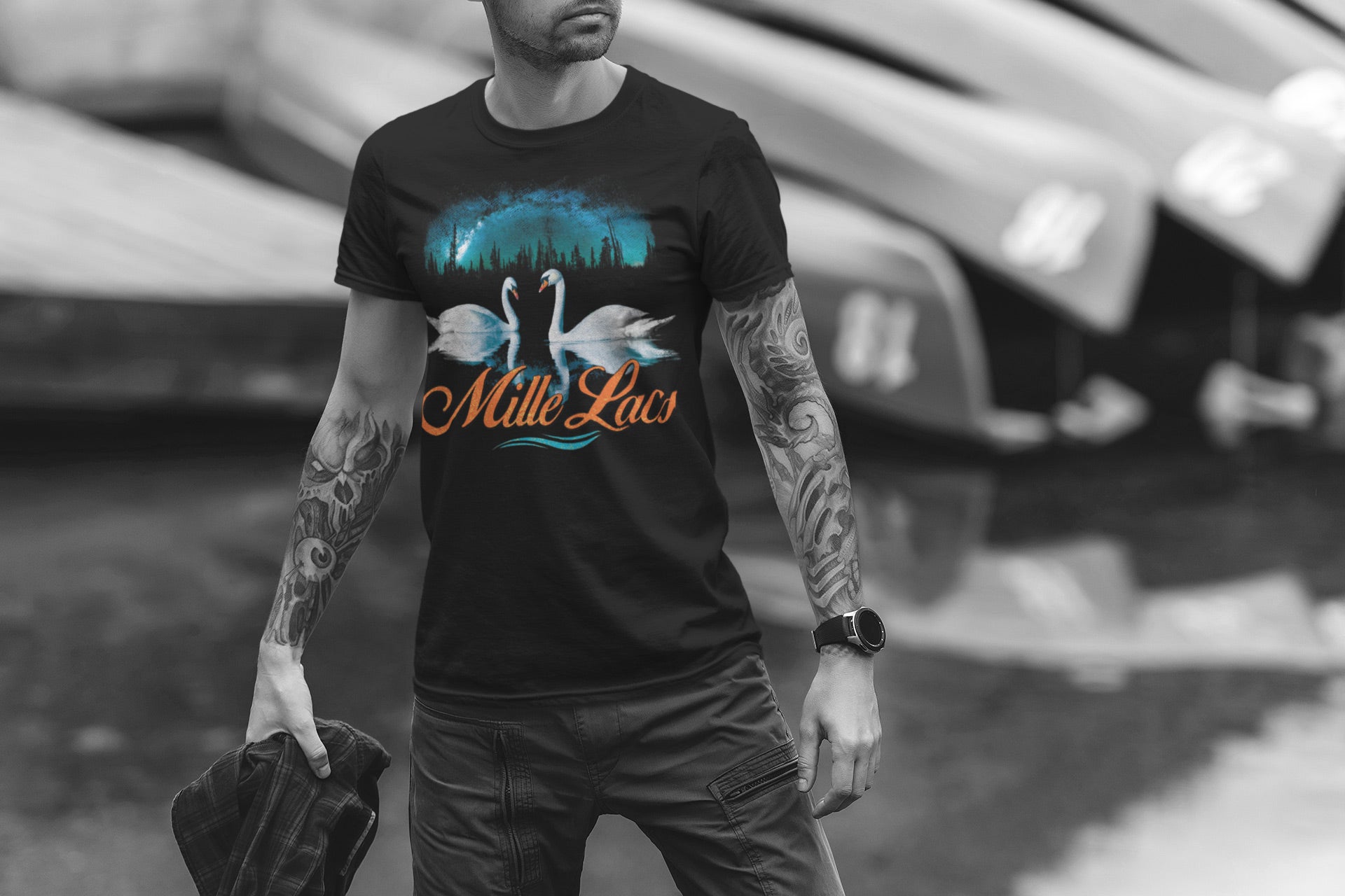 Man with tattoos sporting a Mille Lacs Lake Minnesota shirt featuring two swans in front of a pine forest and the northern lights.