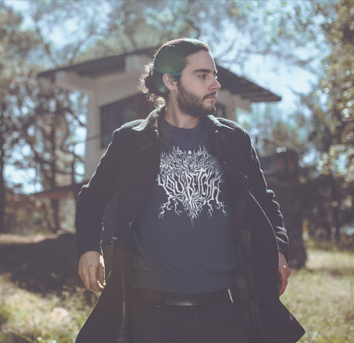 Man with a crew neck sweatshirt with a "You Betcha" metal-band-inspired logo underneath a black peacoat, while standing in front of a cabin on Lake Superior.