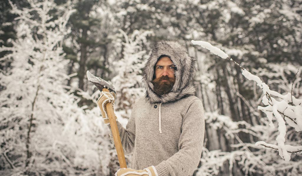 A man in a merino wool sweater, deerskin gloves and fur hat about to chop wood. He stands in a snowy pine forest in Northern Minnesota.