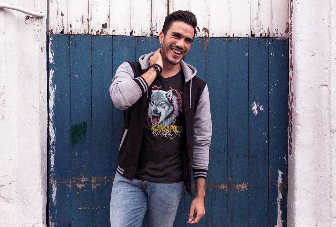 Man standing in front of a workshop smiling and rubbing his neck, while wearing a black-and-gray letterman's jacket over a black graphic tee depicting a wolf surrounding by red lighting and text that reads, "Minneapolis, Minnesota"