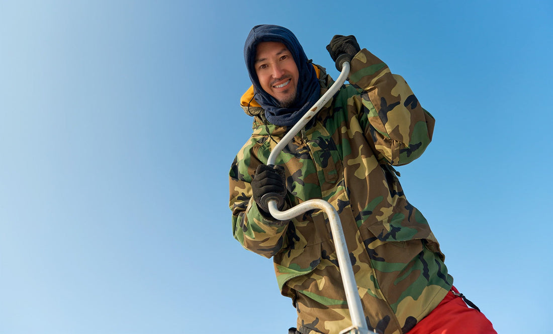 A man drilling a hole for ice fishing in northern minnesota. He wears a camo jacket and orange snowpants and dark hood and gloves.